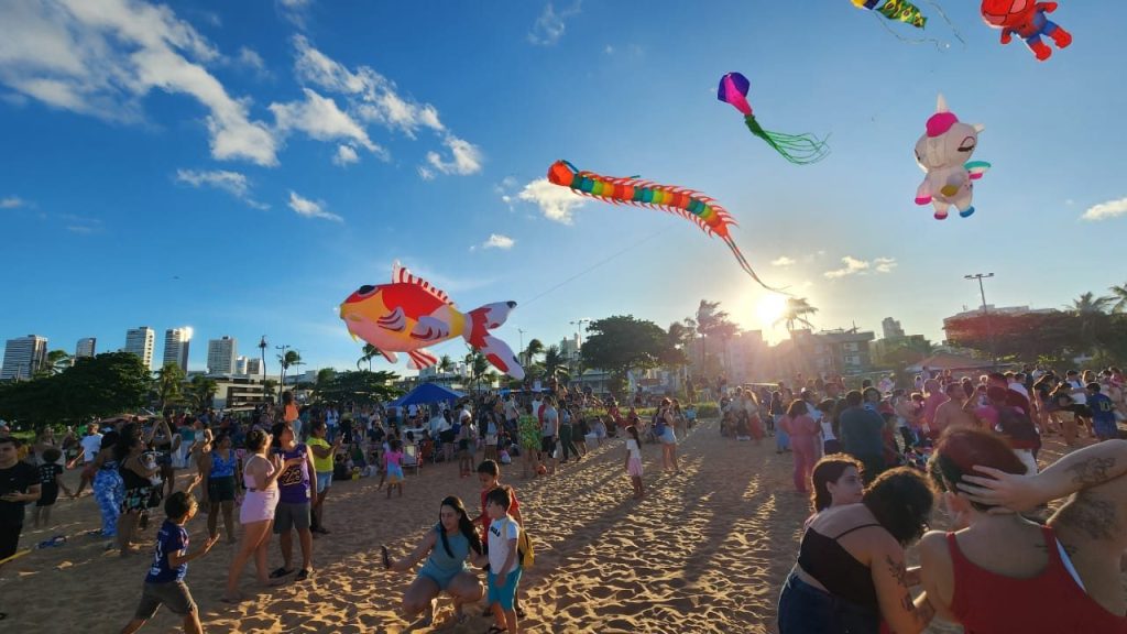 Público na praia ao pôr do sol com pipas gigantes no céu